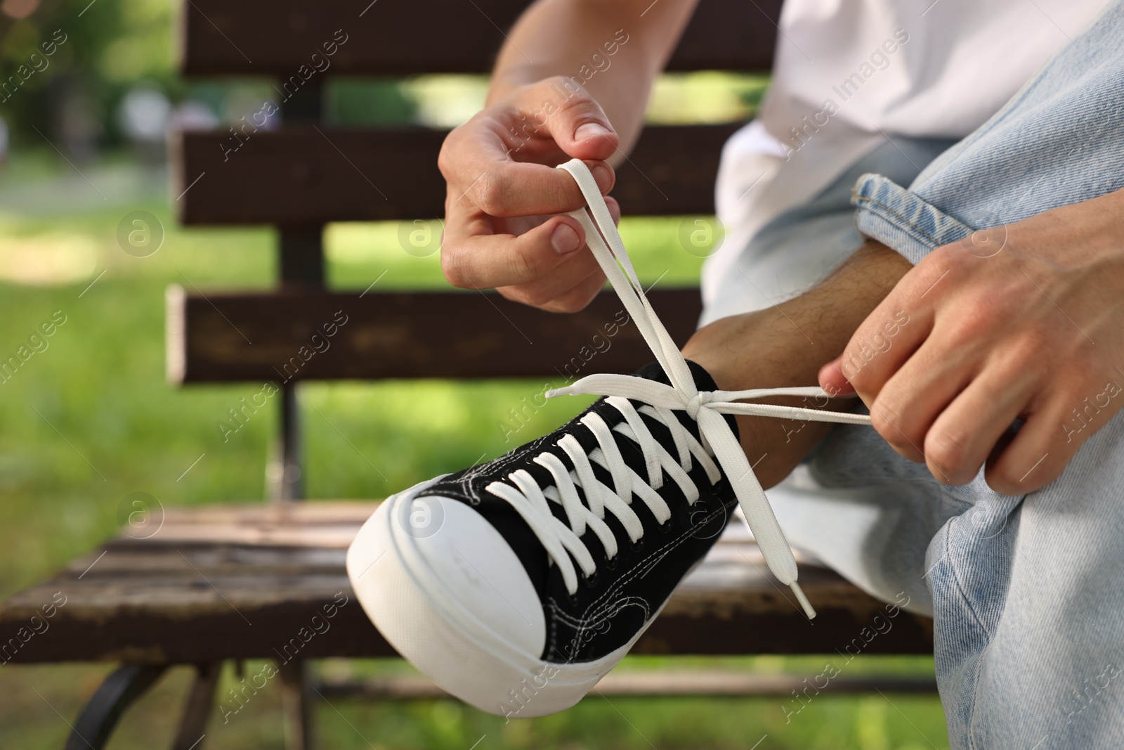 Photo of Man tying shoelace of black sneaker on bench outdoors, closeup