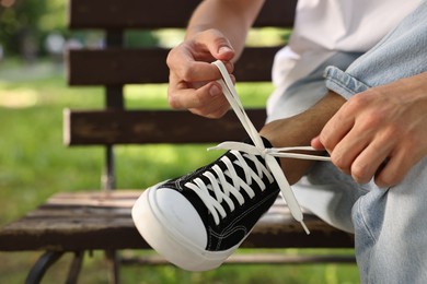 Photo of Man tying shoelace of black sneaker on bench outdoors, closeup