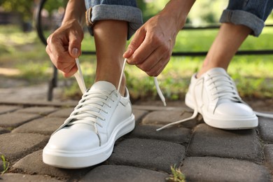 Photo of Woman tying shoelace of white sneaker outdoors, closeup