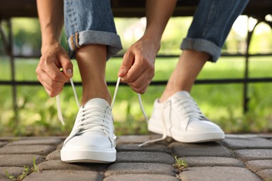 Photo of Woman tying shoelace of white sneaker outdoors, closeup