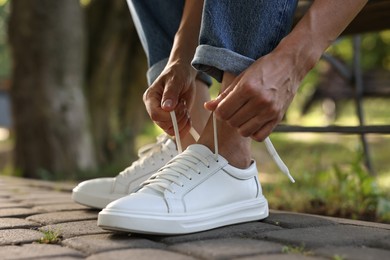 Photo of Woman tying shoelace of white sneaker outdoors, closeup