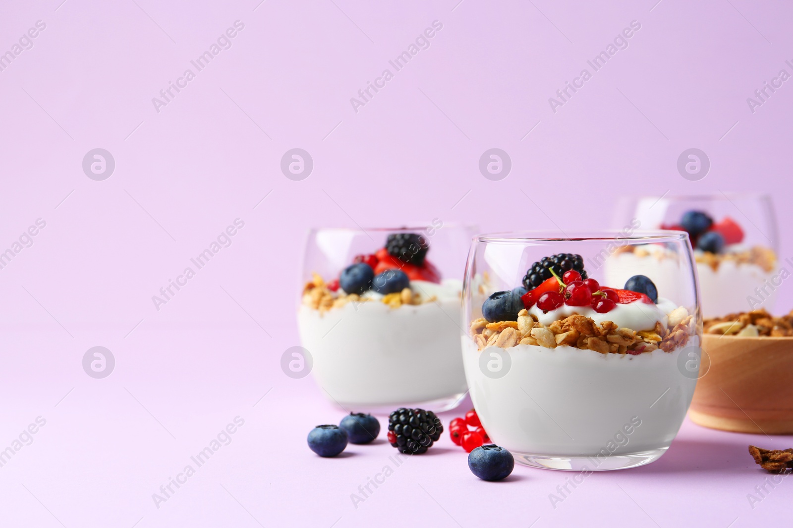 Photo of Tasty yogurt with fresh berries and granola in glasses on violet background, space for text