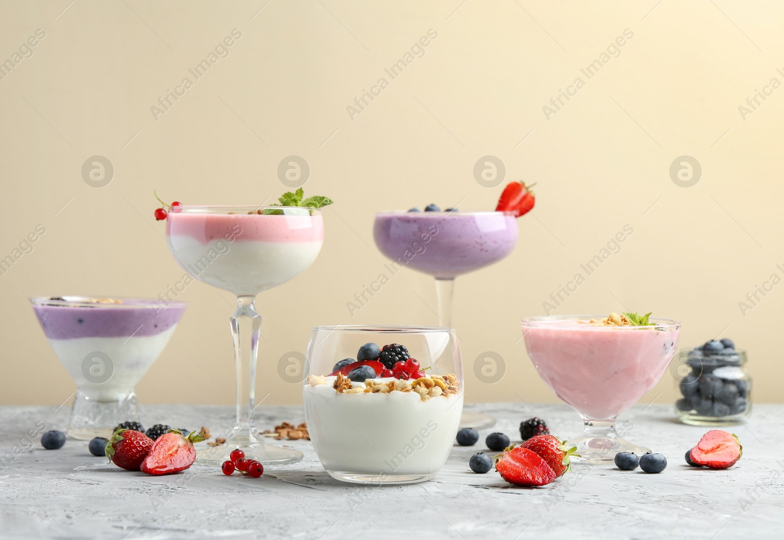 Photo of Different tasty yogurts with fresh berries and granola in glass dishware on gray textured table against pale yellow background