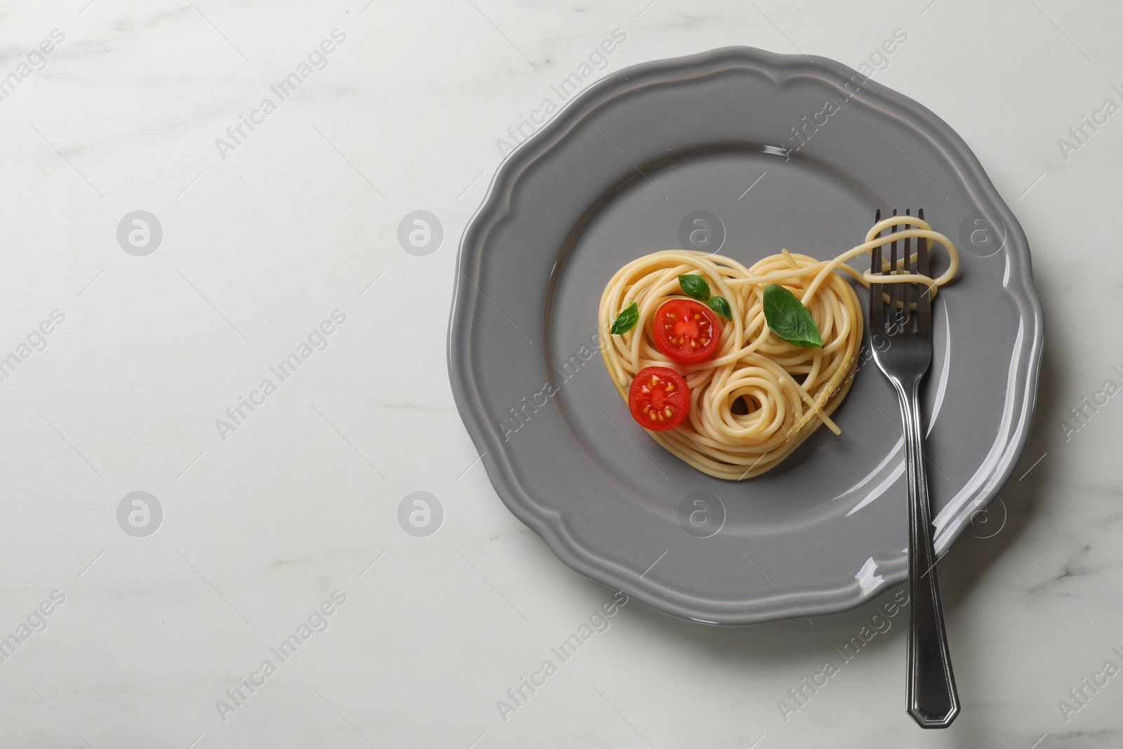 Photo of Heart made of tasty spaghetti, fork, tomato and basil on white marble table, top view. Space for text