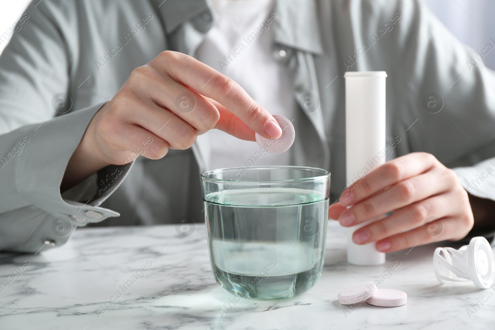 Photo of Woman putting effervescent pill into glass of water at white marble table, closeup