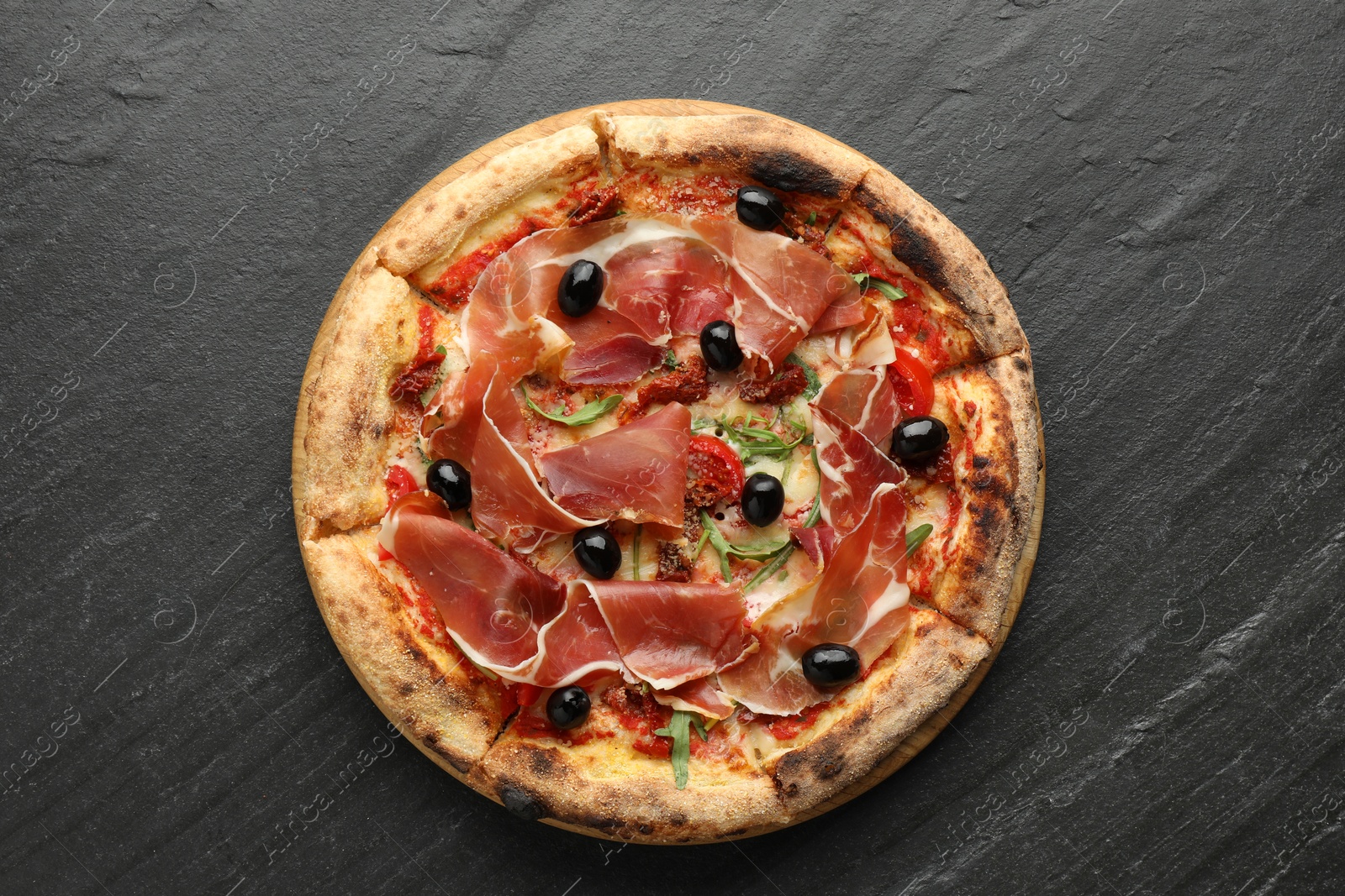 Photo of Tasty pizza with cured ham, olives and sun-dried tomato on black table, top view