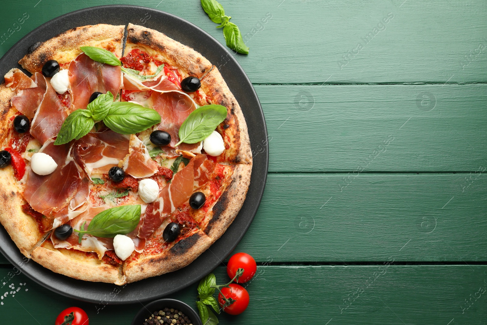 Photo of Tasty pizza with cured ham, olives, mozzarella cheese, tomatoes and arugula on green wooden table, flat lay. Space for text