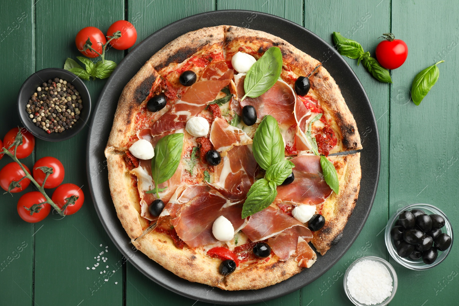 Photo of Tasty pizza with cured ham, olives, mozzarella cheese, tomatoes and arugula on green wooden table, flat lay