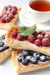 Tasty puff pastries with berries and tea on white wooden table, closeup