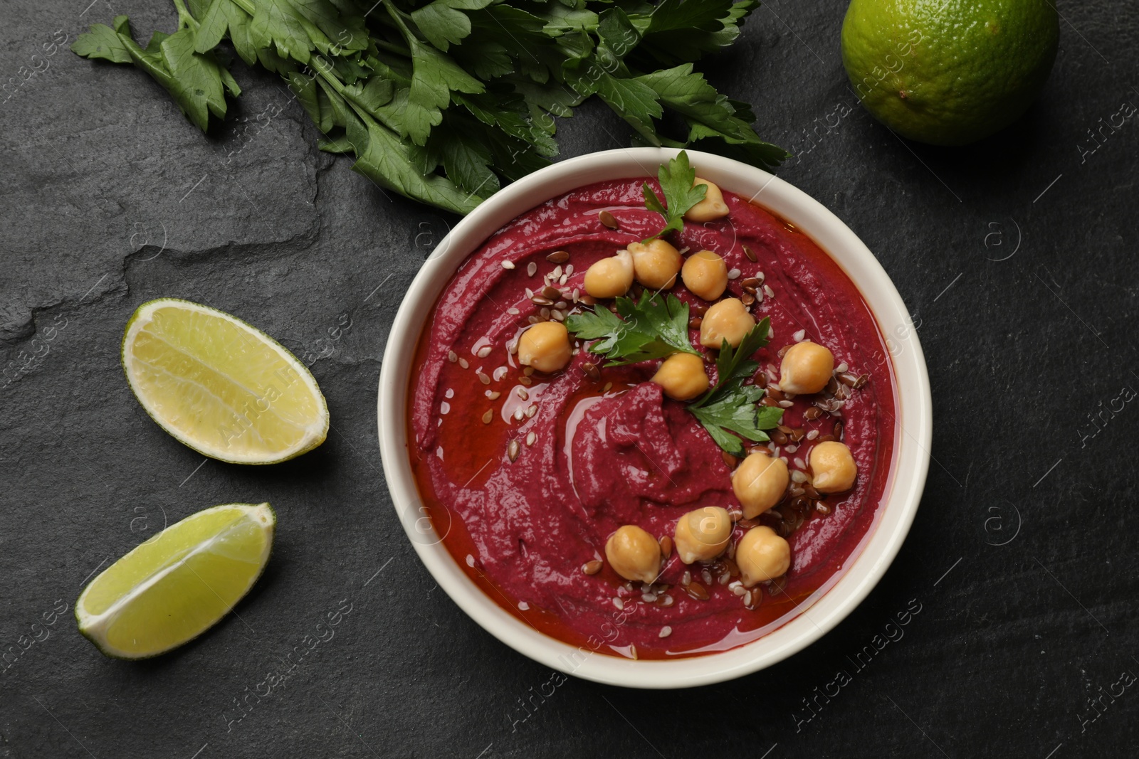 Photo of Tasty beet hummus with chickpeas in bowl, lime and parsley on grey textured table, flat lay