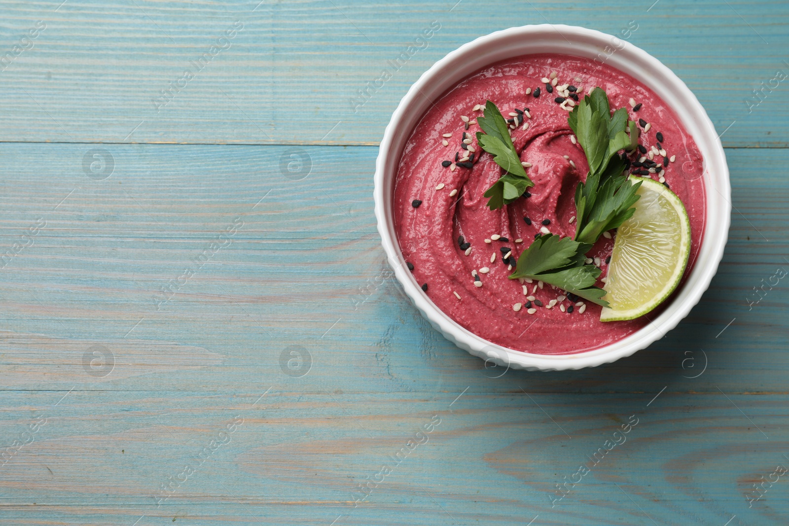 Photo of Tasty beet hummus with parsley and lime in bowl on light blue wooden table, top view. Space for text