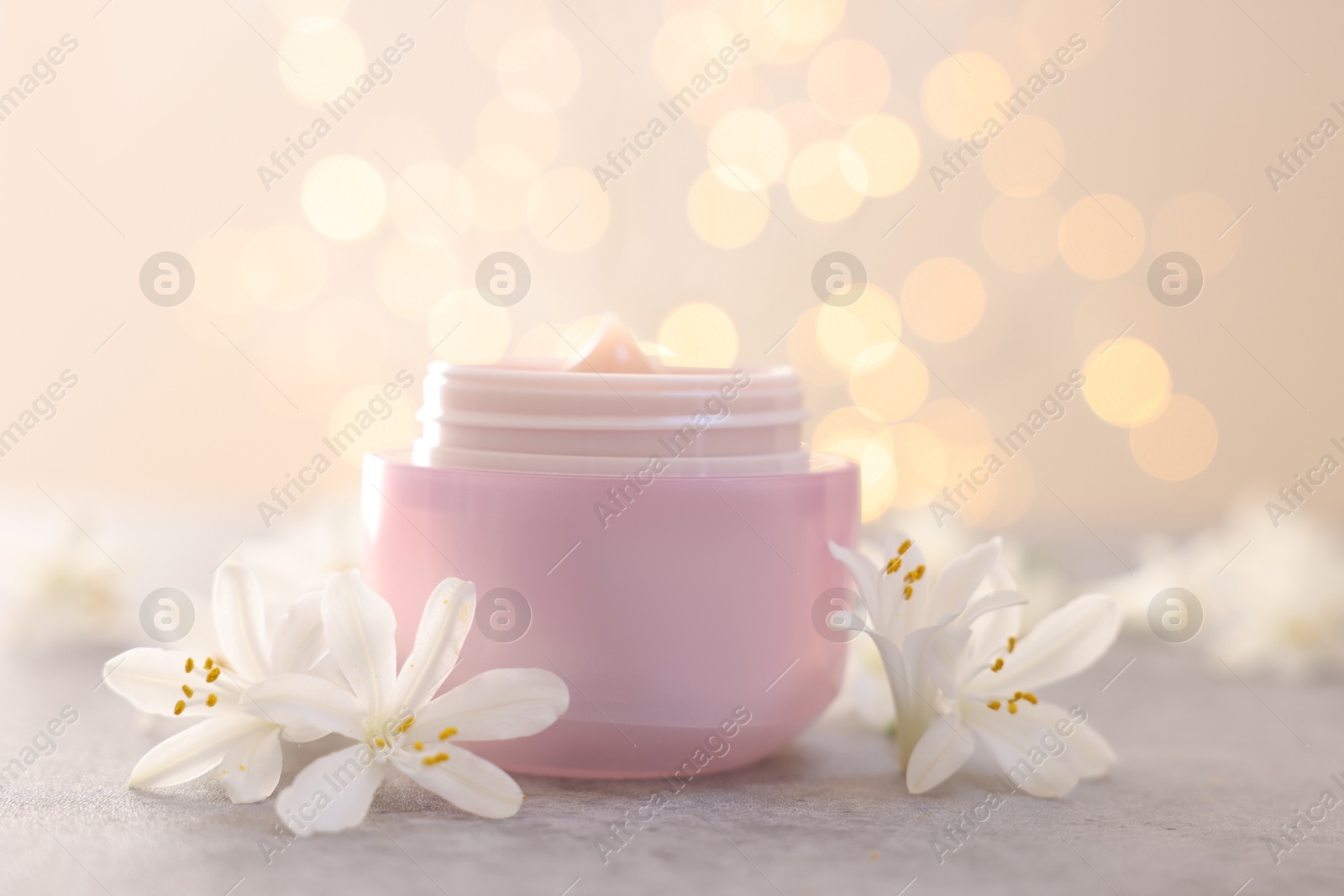 Photo of Cream in jar and jasmine flowers on grey table against beige background with blurred lights, closeup