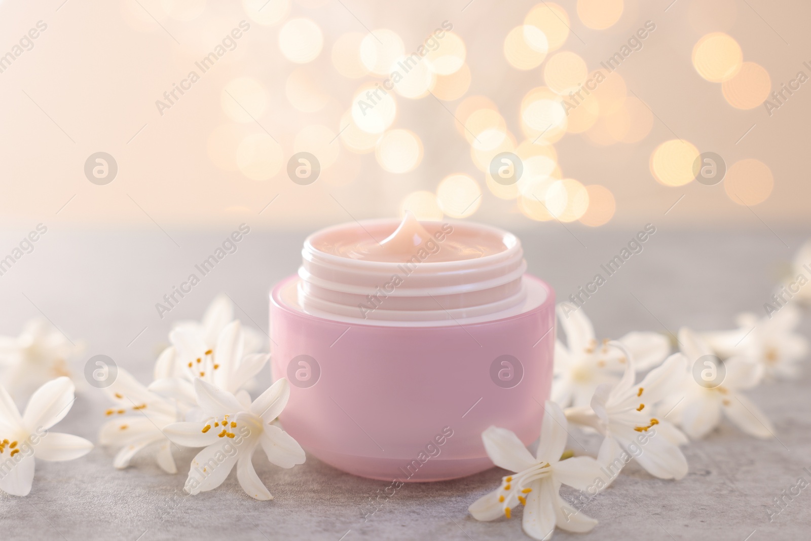 Photo of Cream in jar and beautiful jasmine flowers on grey table against light background with blurred lights, closeup