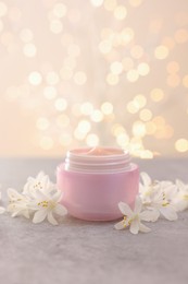 Photo of Cream in jar and beautiful jasmine flowers on grey table against beige background with blurred lights