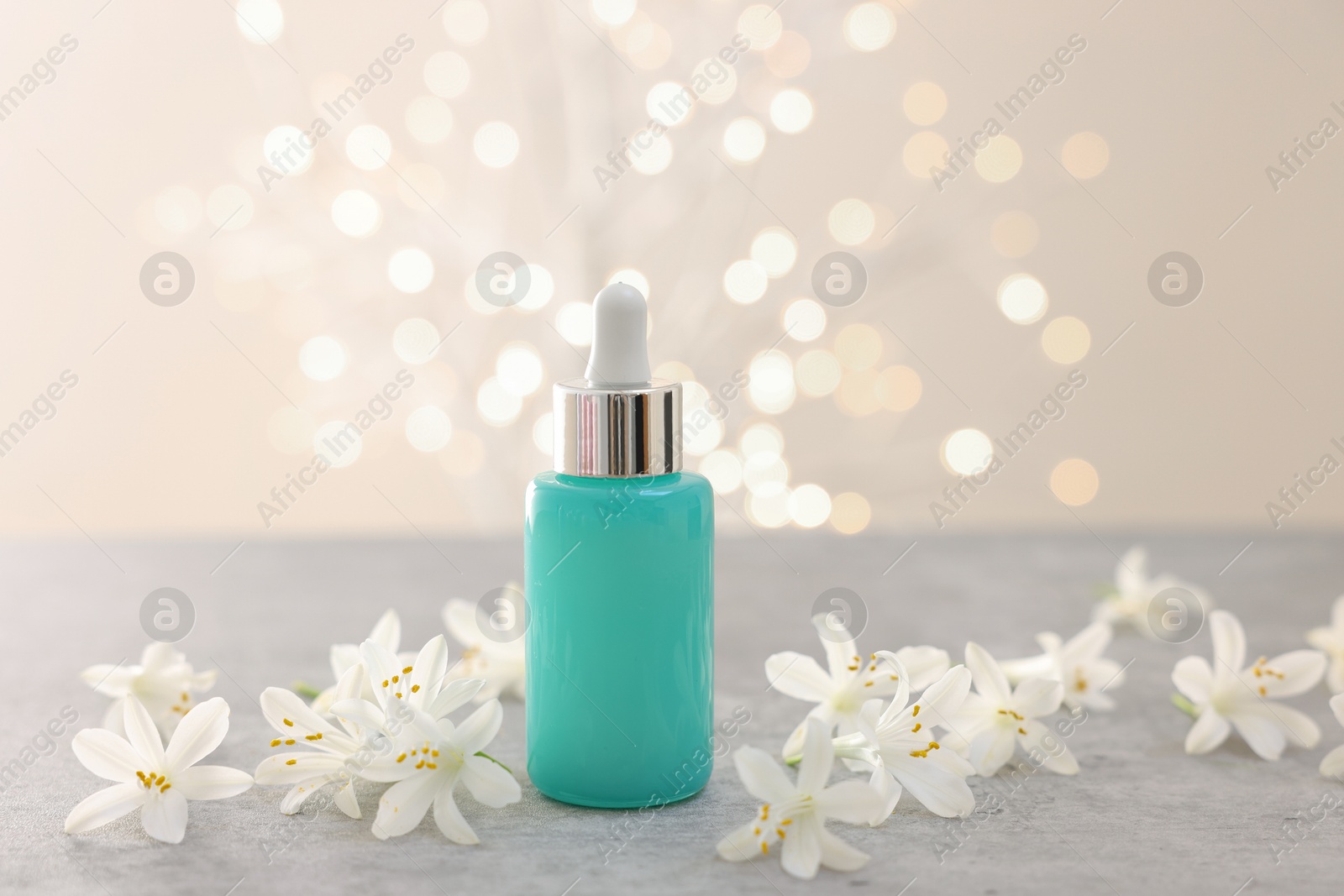 Photo of Cosmetic product in bottle and beautiful jasmine flowers on grey table against beige background with blurred lights, closeup