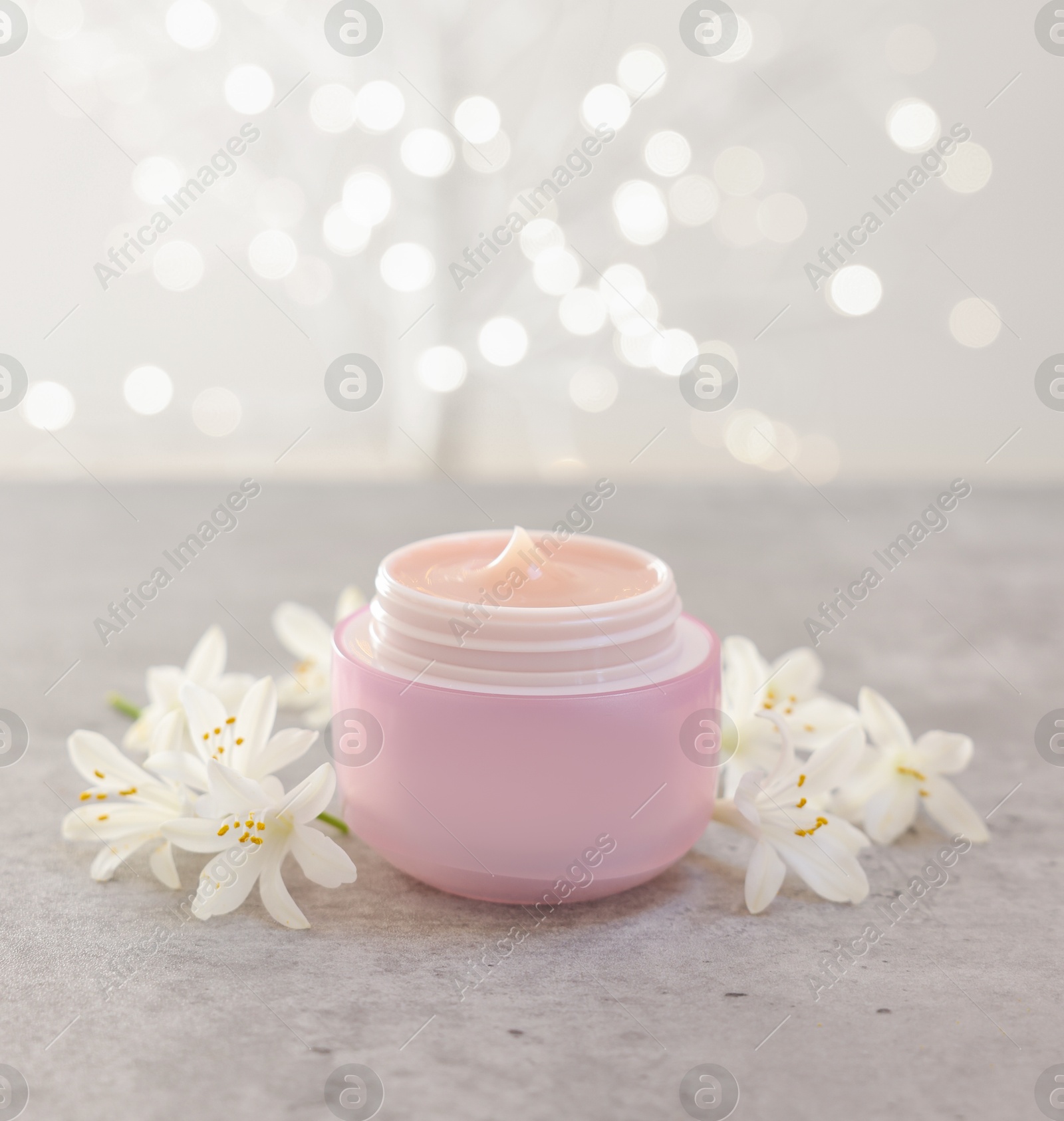 Photo of Cream in jar and beautiful jasmine flowers on grey table against light background with blurred lights, closeup