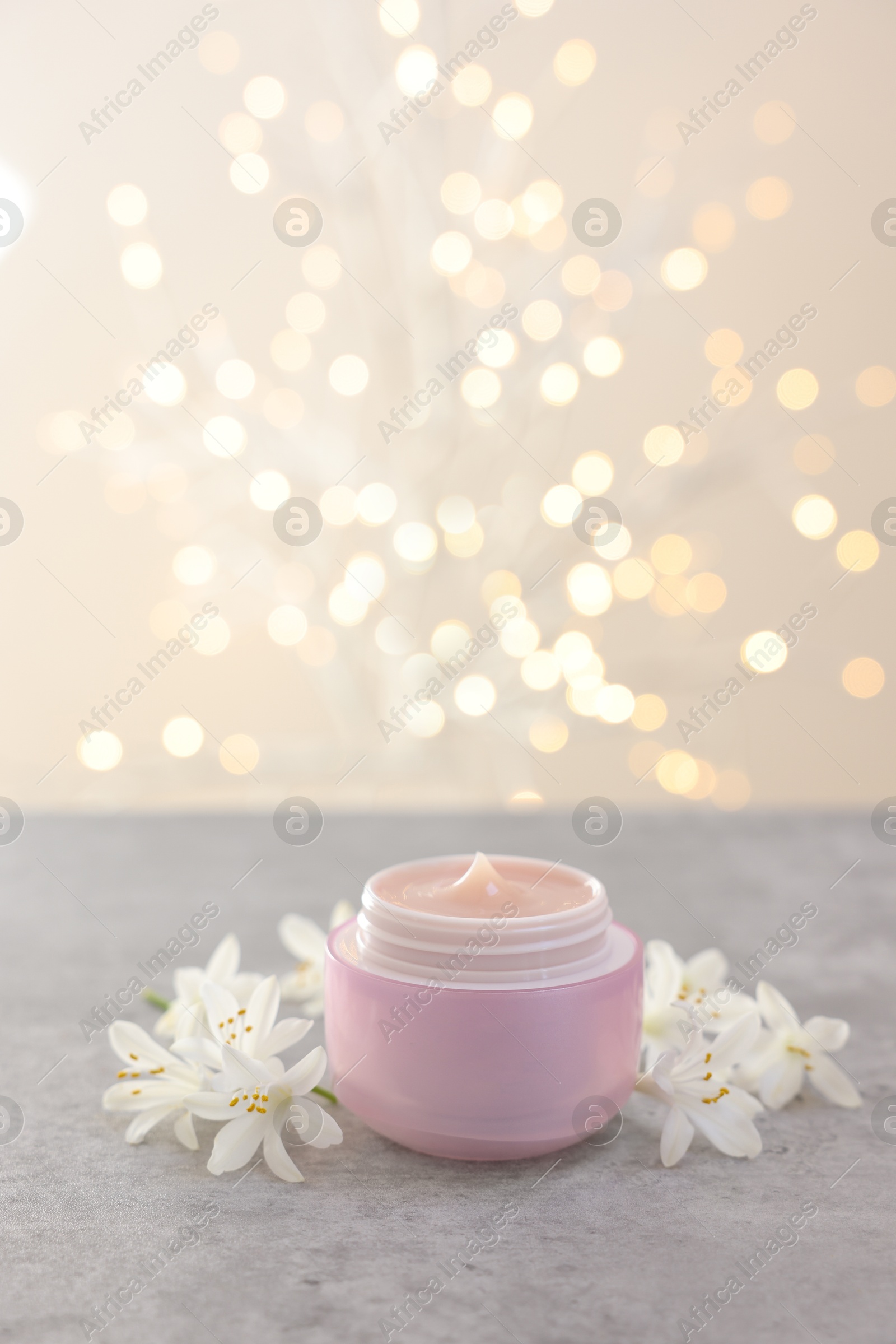 Photo of Cream in jar and beautiful jasmine flowers on grey table against light background with blurred lights, closeup