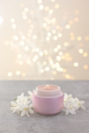 Photo of Cream in jar and beautiful jasmine flowers on grey table against light background with blurred lights, closeup