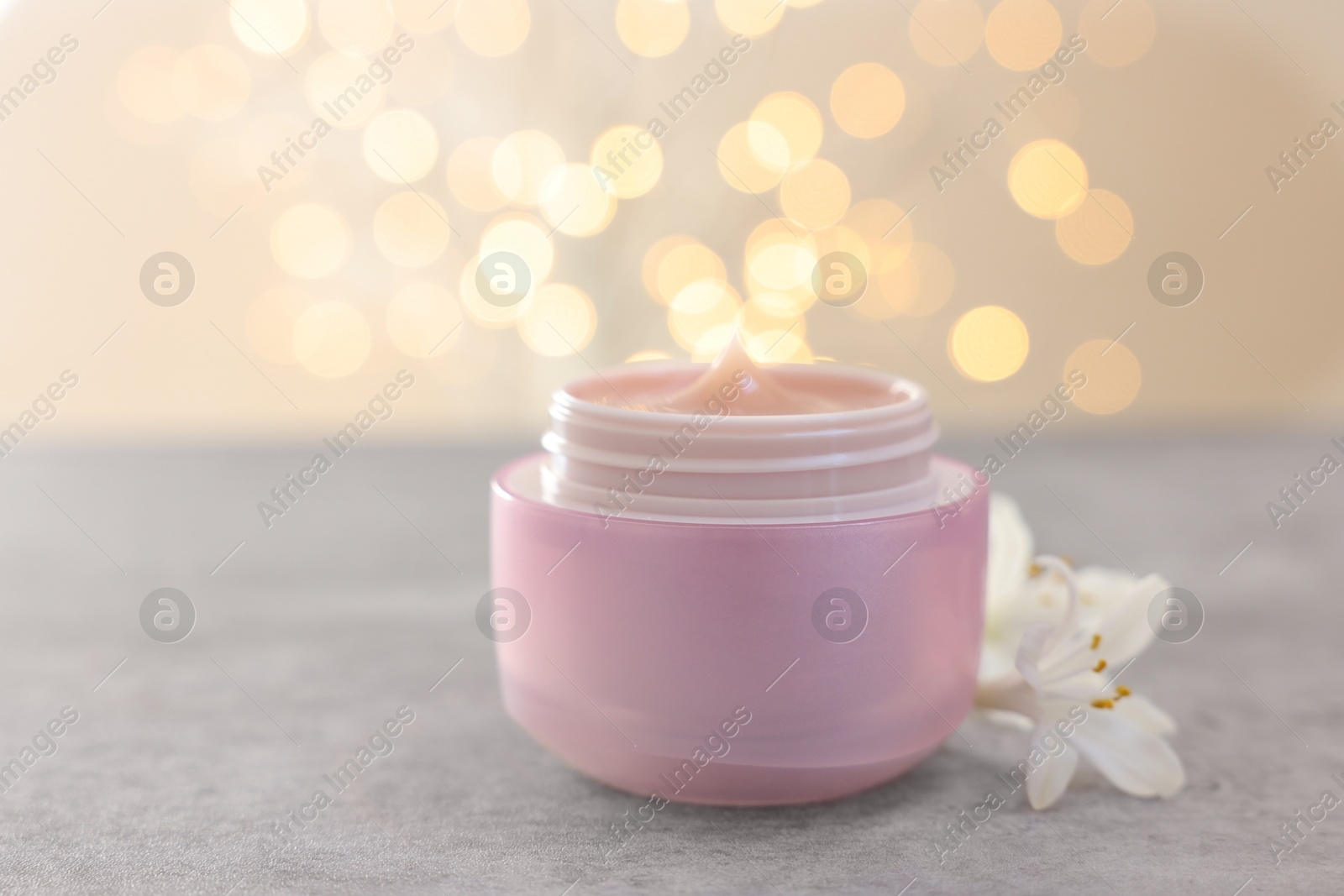 Photo of Cream in jar and beautiful jasmine flowers on grey table against light background with blurred lights, closeup