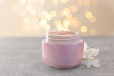 Photo of Cream in jar and beautiful jasmine flowers on grey table against light background with blurred lights, closeup