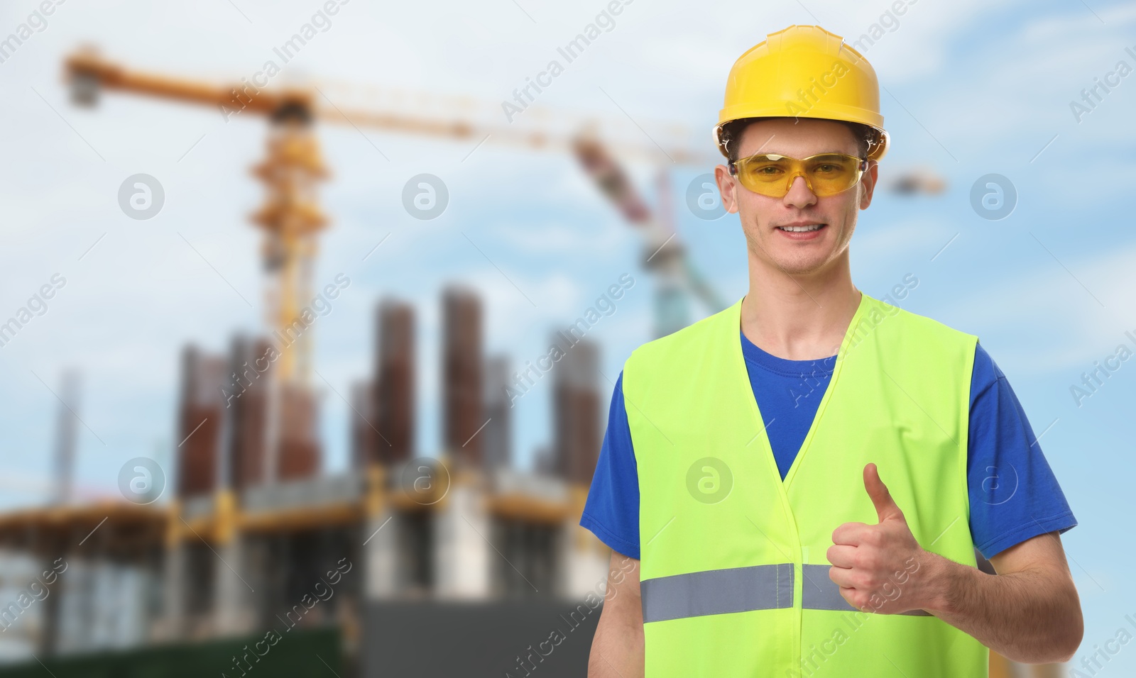 Image of Young man wearing safety equipment and showing thumbs up at construction site. Space for text