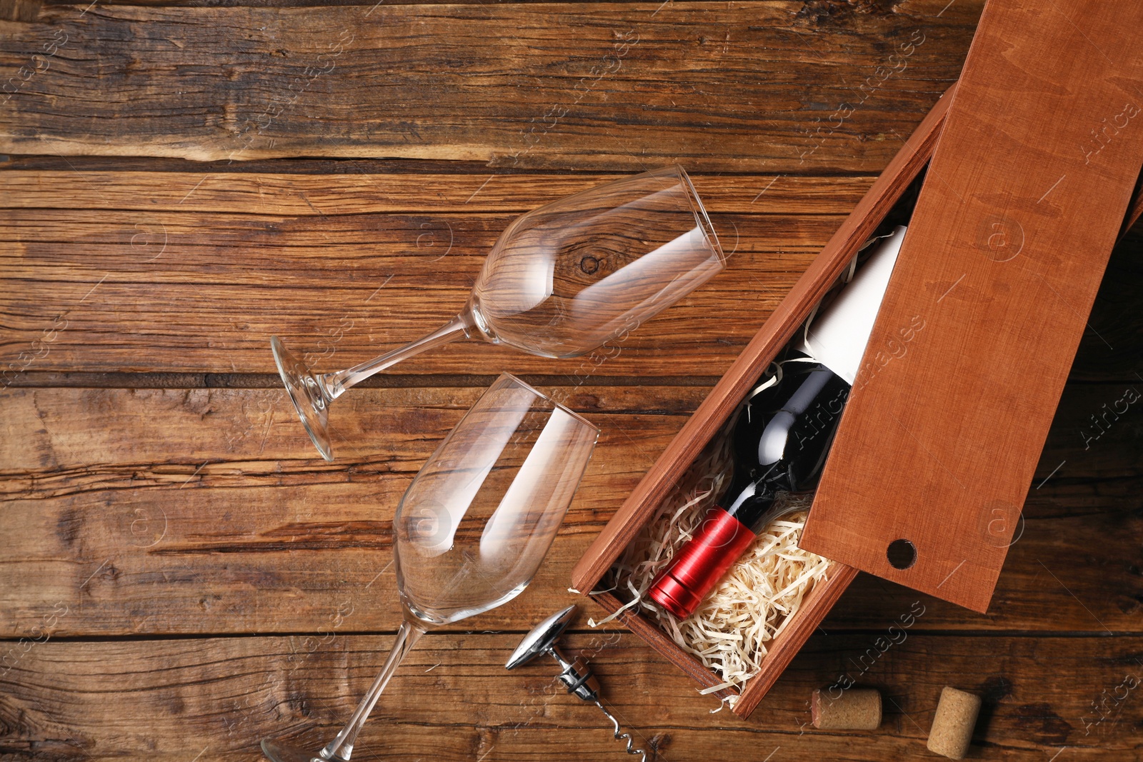 Photo of Box with wine bottle, glasses, corkscrew and corks on wooden table, flat lay