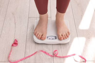 Photo of Woman standing on floor scale and measuring tape at home, closeup. Weight control