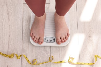 Photo of Woman standing on floor scale and measuring tape at home, top view. Weight control