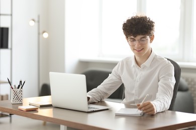 Photo of Teenager taking notes while working with laptop at table indoors. Remote job