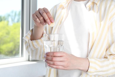 Photo of Woman putting effervescent pill into glass of water indoors, closeup