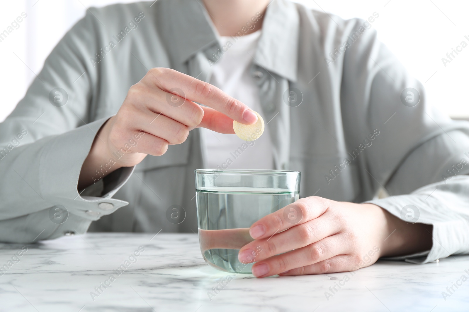 Photo of Woman putting effervescent pill into glass of water at white marble table, closeup