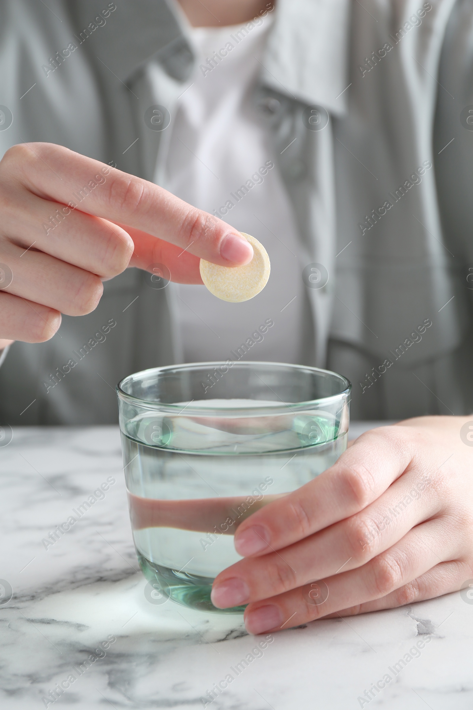 Photo of Woman putting effervescent pill into glass of water at white marble table, closeup