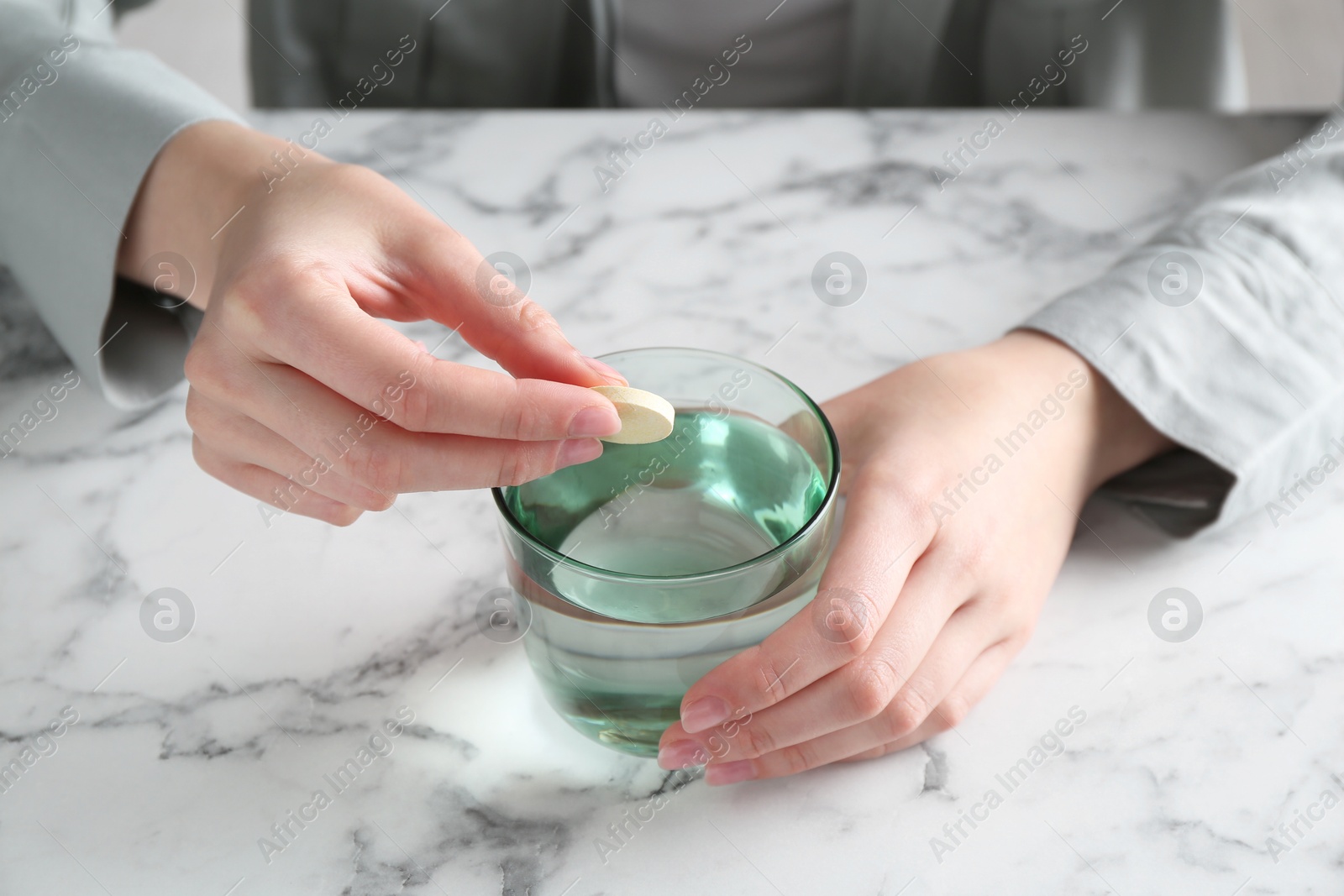 Photo of Woman putting effervescent pill into glass of water at white marble table, closeup