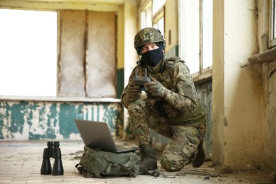 Photo of Military mission. Soldier in uniform using laptop and binoculars inside abandoned building