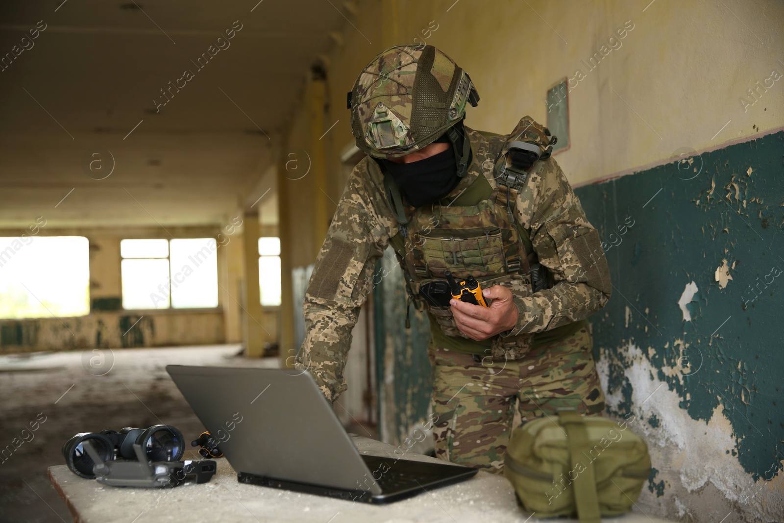 Photo of Military mission. Soldier in uniform with radio transmitter using laptop at table inside abandoned building