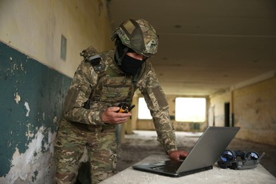 Military mission. Soldier in uniform with radio transmitter using laptop at table inside abandoned building