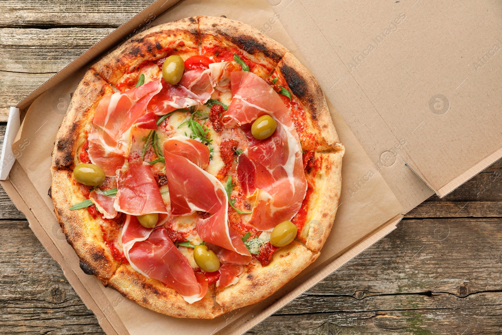 Photo of Tasty pizza with cured ham, olives and sun-dried tomato in cardboard box on wooden table, top view