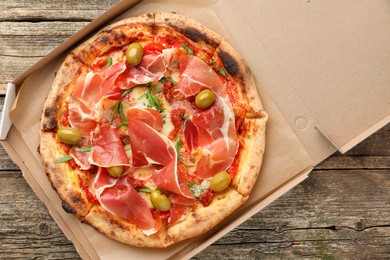 Tasty pizza with cured ham, olives and sun-dried tomato in cardboard box on wooden table, top view