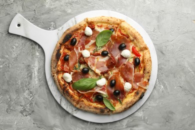 Photo of Tasty pizza with cured ham, olives, sun-dried tomato and basil on gray textured table, top view