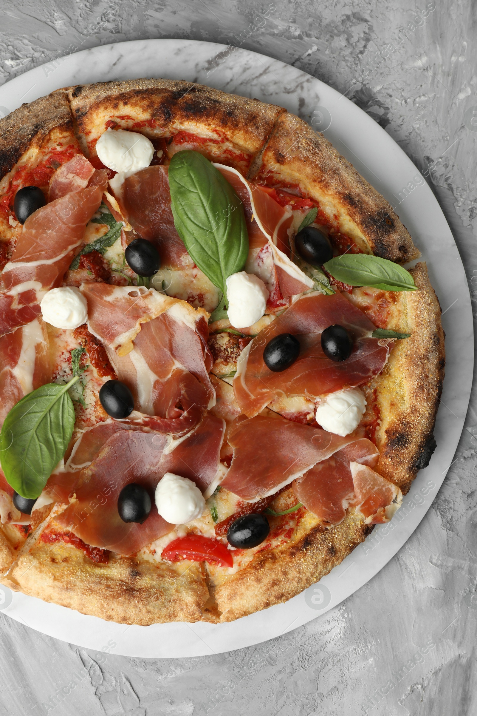 Photo of Tasty pizza with cured ham, olives, sun-dried tomato and basil on gray textured table, top view