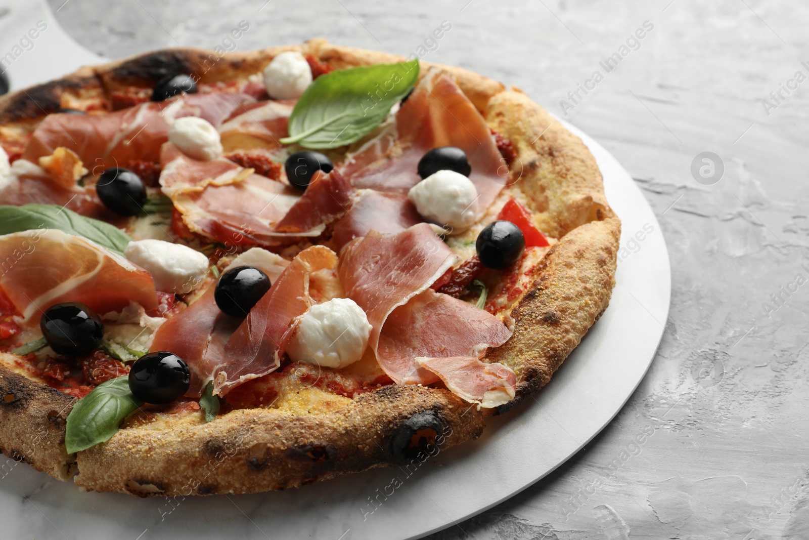 Photo of Tasty pizza with cured ham, olives, mozzarella cheese, sun-dried tomato and basil on gray textured table, closeup