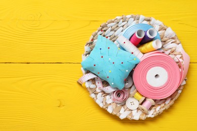 Light blue pincushion, pins and other sewing tools on yellow wooden table, top view. Space for text