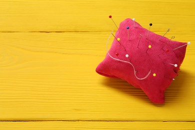 Red pincushion with sewing pins on yellow wooden table. Space for text