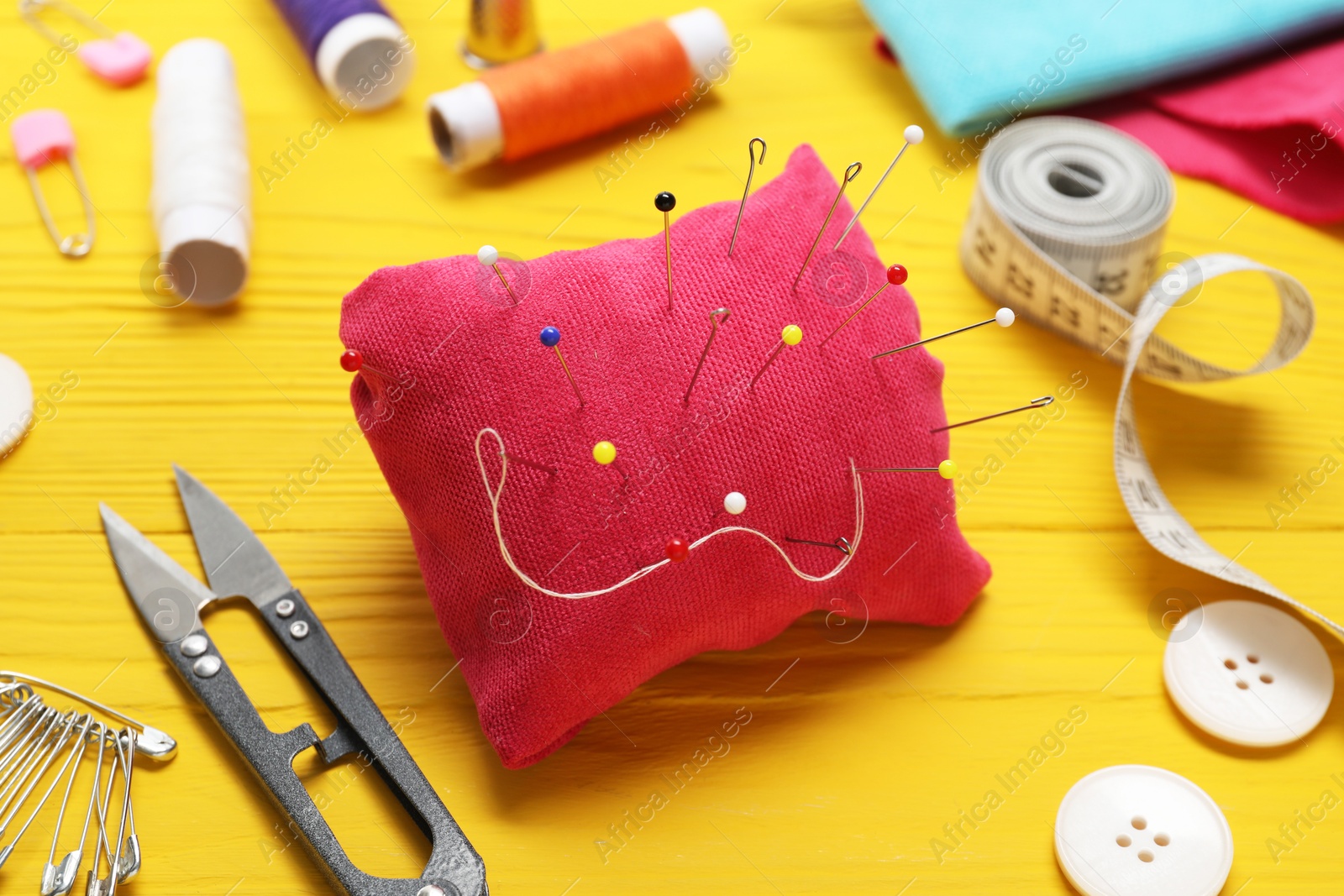 Photo of Red pincushion with pins and other sewing tools on yellow wooden table, closeup