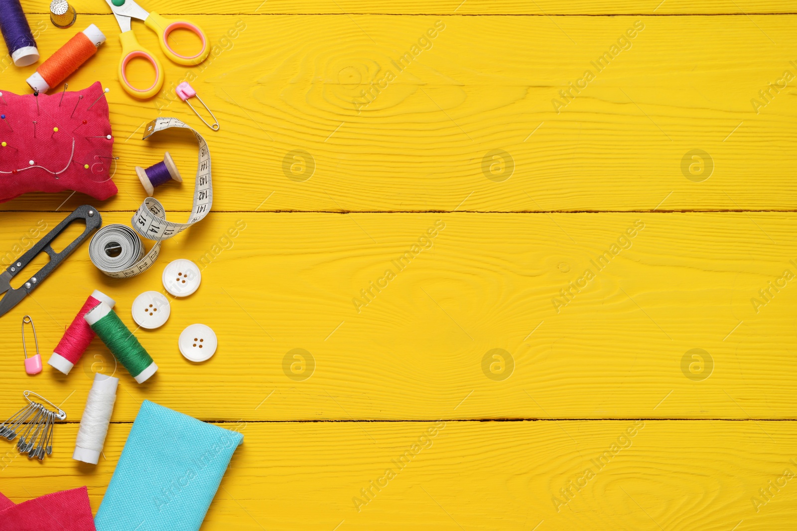 Photo of Red pincushion with pins and other sewing tools on yellow wooden table, flat lay. Space for text