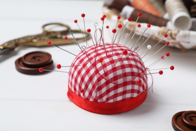 Checkered pincushion with pins and other sewing tools on white wooden table, closeup