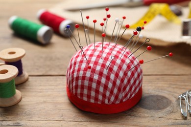 Photo of Checkered pincushion with pins and other sewing tools on wooden table, closeup