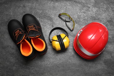 Pair of working boots, hard hat, protective goggles and earmuffs on grey surface, top view