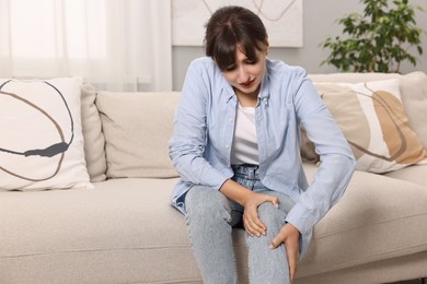 Upset woman suffering from knee pain on sofa at home