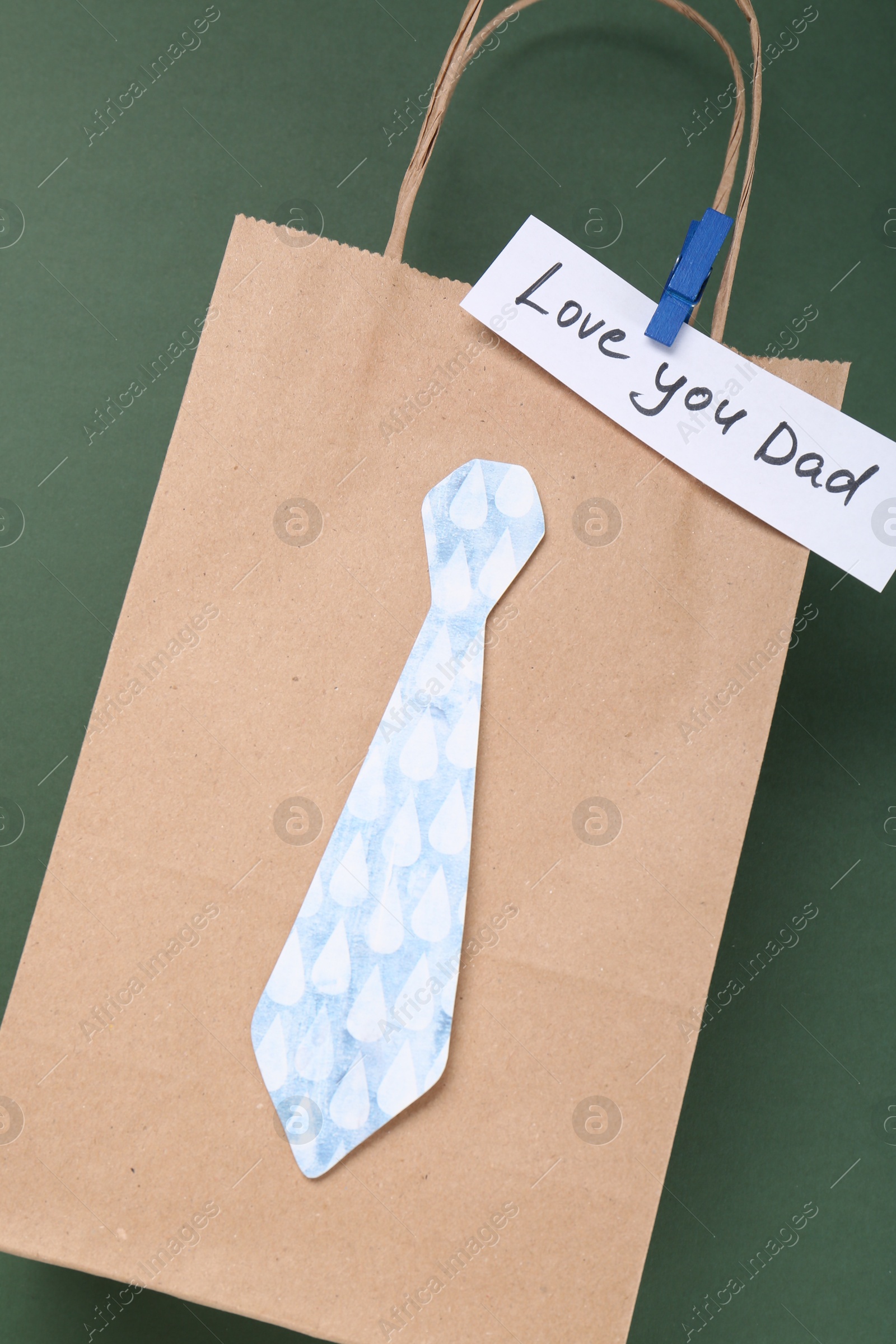 Photo of Happy Father's Day. Paper bag with tie and phrase Love You Dad on green background, top view